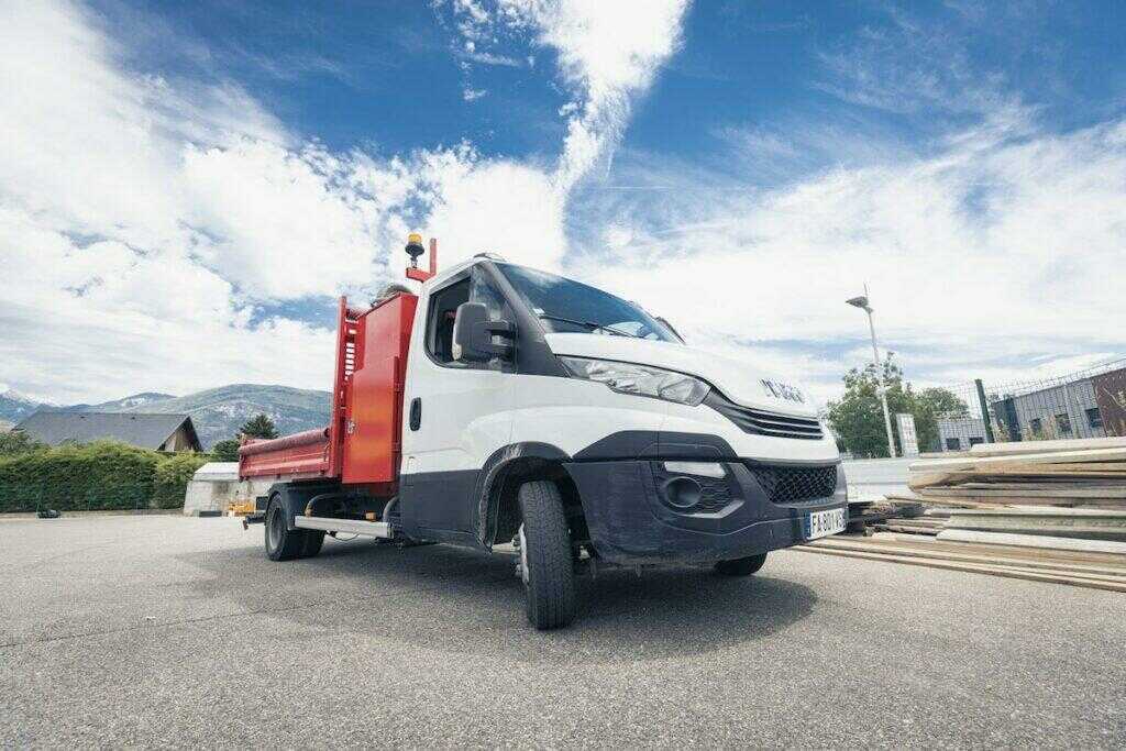 Photo avec, au centre un camion garé à côté d'une pile de planches en bois, et un ciel bleu de fond
